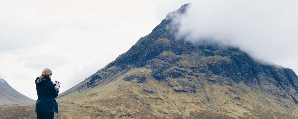 A woman stands with her camera pointing towards a cloud-shrouded mountain in Glen Coe, Scotland